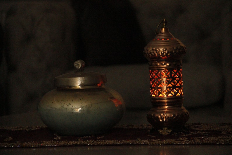 May include: A copper-colored lantern with a geometric pattern and a small, round, lidded jar. The lantern is lit from within, casting a warm glow. The jar is a muted teal color with a silver lid.