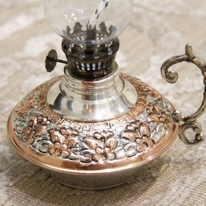 Pode incluir: Uma l&acirc;mpada de &oacute;leo de prata e cobre com um design floral ornamentado e uma chamin&eacute; de vidro. A l&acirc;mpada tem uma al&ccedil;a e uma pequena ma&ccedil;aneta no lado.