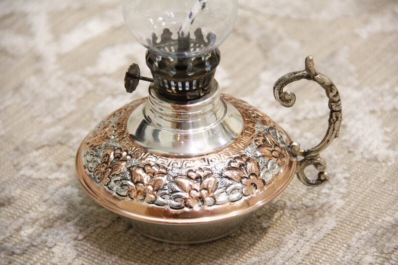 Pode incluir: Uma l&acirc;mpada de &oacute;leo de prata e cobre com um design floral ornamentado e uma chamin&eacute; de vidro. A l&acirc;mpada tem uma al&ccedil;a e uma pequena ma&ccedil;aneta no lado.