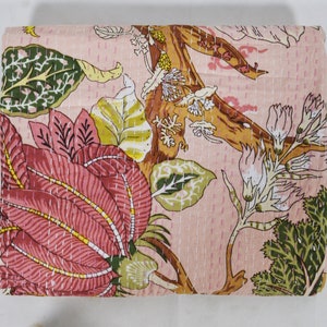 Könnte beinhalten: Eine gefaltete Decke mit floralem Muster. Das Design zeigt eine große, stilisierte Blume in Rosa und Rot, mit grünen und gelben Blättern und braunen Ästen auf einem hellrosa Hintergrund. Weiße Nähte sind über die gesamte Decke sichtbar.