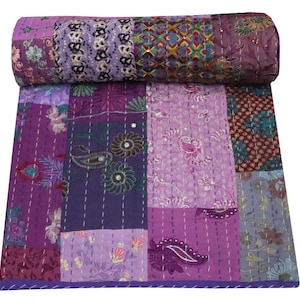 May include: A patchwork quilt in shades of purple, featuring various floral and paisley patterns. The quilt is made of different fabric squares stitched together with white thread. Rolled up at the top, it displays a variety of textures and designs.