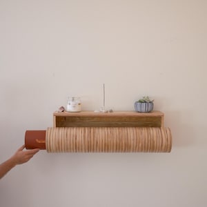 May include: A modern, wall-mounted shelf made of light-colored wood. The shelf has a cylindrical, ribbed design and a rectangular top shelf. A rolled-up brown item is partially pulled out. A small succulent in a blue and white pot sits on the top shelf.