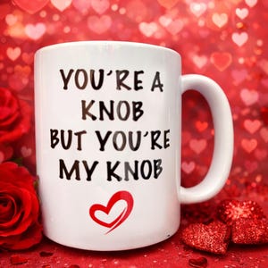 May include: White ceramic mug with the text "YOU'RE A KNOB BUT YOU'RE MY KNOB" in black, and a red heart graphic. The mug is set against a red background with heart-shaped bokeh and a red rose.