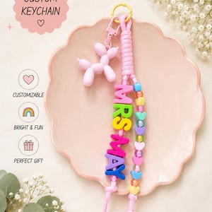 May include: A custom keychain featuring a pink balloon dog charm, a braided pink cord, and colorful letter beads spelling MRS MAY. The keychain includes a strand of multi-colored heart-shaped beads and a yellow ring attachment. Text includes CUSTOM KEYCHAIN, CUSTOMIZABLE, BRIGHT & FUN, PERFECT GIFT, MADE WITH CARE, and MADE FOR YOU.