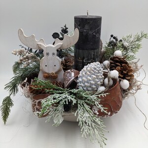 May include: A decorative centerpiece featuring a white moose figurine, a black pillar candle, pine cones, and faux greenery. The arrangement is set in a rustic, brown-edged container, creating a festive, wintery aesthetic. The candle has the text "Wax Country".