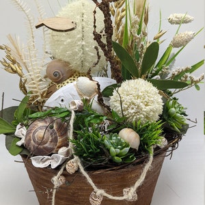 以下が含まれることがあります： 風化した金属製のポットに入った素朴な装飾アレンジメント。木製の魚、貝殻、白い魚、さまざまな乾燥植物と草が特徴です。アレンジメントには十字架と貝殻のひもが含まれています。