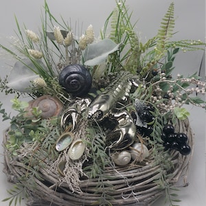 以下が含まれることがあります： 緑、貝殻、銀色のロブスターの置物が入った、織りバスケットが特徴の装飾的なセンターピース。濃い灰色のカタツムリの殻と小さな黒いベリーがアクセントになっています。アレンジメントは白い背景に設定されています。