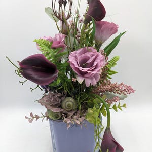Könnte beinhalten: Blumenarrangement in einer quadratischen, hellvioletten Glasvase. Die Anordnung umfasst dunkelviolette Calla-Lilien, rosa Rosen und verschiedene grüne Blätter. Die Gesamtästhetik ist elegant und dekorativ.