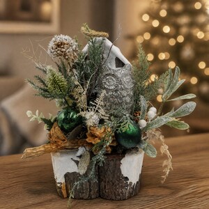 Kerstarrangement met uil, kerstballen, dennenappels, groen en zilver, in een pot van boomschors.