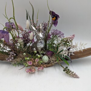 May include: An Easter-themed centerpiece featuring a silver bunny figurine surrounded by purple and white flowers, green foliage, and decorative eggs. The arrangement is set in a long, wooden, boat-shaped container, creating a festive display.