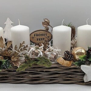 May include: A woven basket arrangement features four white pillar candles, festive decorations, and foliage. The display includes a wooden sign with the text "Frohes Fest". The centerpiece is decorated with gold and rose-gold accents, and a white star.