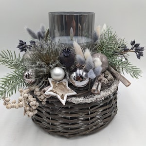 May include: A decorative centerpiece featuring a glass candle holder surrounded by dried flowers, silver ornaments, and a star-shaped accent. The arrangement sits in a woven basket, creating a rustic and festive look. The color palette includes silver, gray, and natural tones.