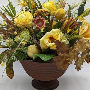 Peut inclure: Composition florale décorative dans un bol marron et côtelé. L'arrangement comprend des roses jaunes, des tournesols et divers feuillages, dont des feuilles et des épis de blé. La palette de couleurs est chaude, avec des nuances de jaune, de marron et de vert.
