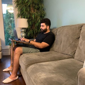 May include: A person using a laptop on a wooden lap desk while seated on a grey sofa. The person wears a black top, green shorts, and white slippers. A lamp and a large plant are in the background.