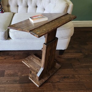Peut inclure: Une table d'appoint en forme de C en bois teinté brun foncé avec un plateau rectangulaire et une base large. La table présente une colonne de support verticale et une surface de plateau horizontale. Un livre à couverture blanche repose dessus.