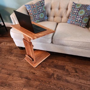 May include: A C-shaped wooden side table with a natural wood grain finish, featuring a rectangular top and a sturdy base. A black laptop is placed on the surface of the table, which is positioned next to a beige tufted sofa with two patterned cushions.