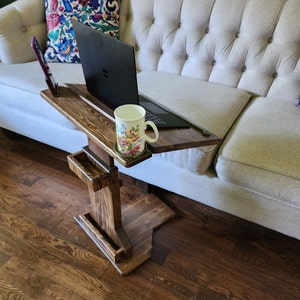 May include: A dark stained wooden laptop table with a built-in phone holder and a small tray. A laptop and a floral mug are on the table. The table is next to a beige couch with decorative pillows.