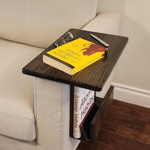 May include: A dark stained wooden side table with a book, glasses, and a pen on top. The book is titled "The Design of Everyday Things." A second book, "Atomic Habits," and a phone are in a lower shelf.