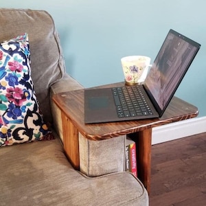 May include: A dark brown wooden side table with a built-in shelf, holding a laptop, a floral teacup, and two books. The table is attached to a grey upholstered armchair, with a decorative floral cushion.