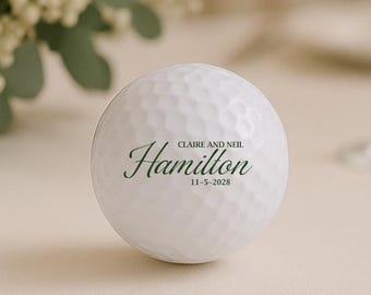 May include: A white golf ball with the name "Hamilton" in a script font, along with the date "11-5-2028". The golf ball is on a light-colored surface, with a blurred background of flowers and a wine glass.