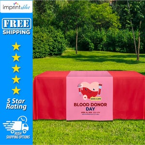 May include: Red table runner with a pink blood donor day design. The design features a cartoon heart with a needle and test tubes, and the text "Save a Life!"  Blood Donor Day, April 25, 2028, 9AM-4PM, Keelingborough City Hall.