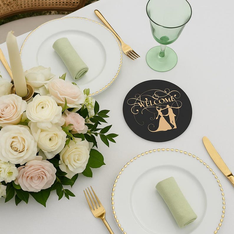 May include: A wedding table setting with white plates, gold cutlery, and a green wine glass. A black coaster with gold text that says "Welcome" and a couple dancing is on the table. A bouquet of white and pink roses and a candle complete the elegant look.
