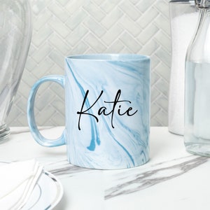 May include: A light blue ceramic mug with a marbled design and the name "Katie" written in black cursive script.