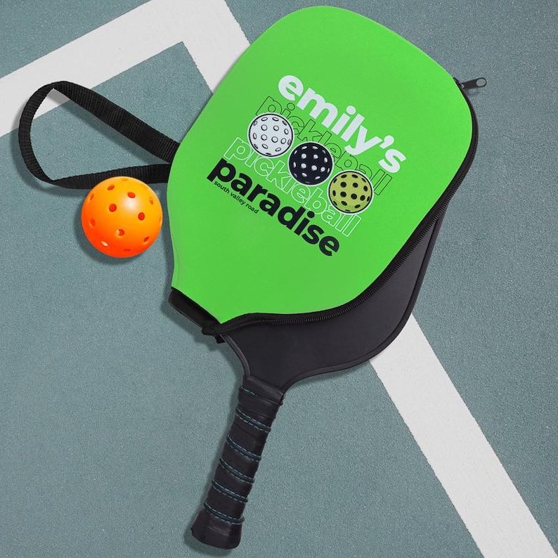 Pode incluir: Uma raquete e capa de pickleball verdes com o texto "emily's pickleball paradise" e tr&ecirc;s gr&aacute;ficos de pickleball. Uma bola de pickleball laranja est&aacute; ao lado da raquete. A raquete tem uma pega preta e uma al&ccedil;a preta.