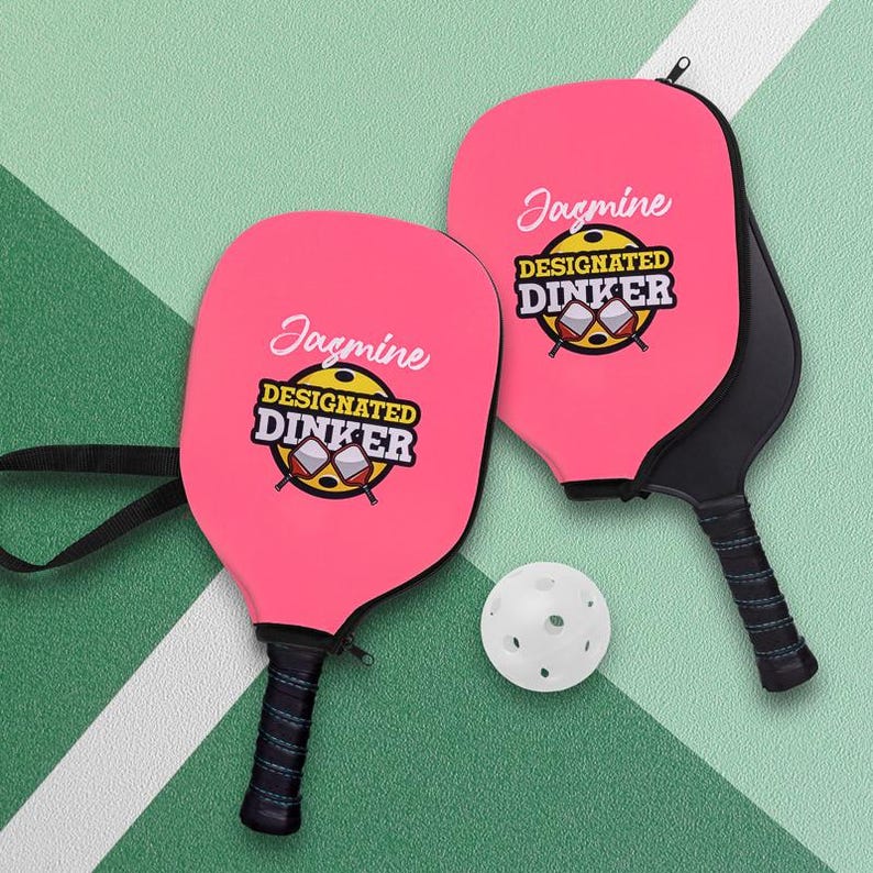 Pode incluir: Duas raquetes de pickleball rosa com o texto "Jasmine DESIGNATED DINKER" e uma bola de pickleball branca em uma quadra verde. As raquetes t&ecirc;m cabos pretos e uma capa com z&iacute;per preta.