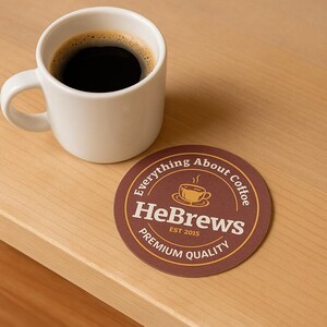 May include: A white ceramic coffee mug filled with black coffee sits next to a brown coaster. The coaster has a circular design with the text "Everything About Coffee" and "HeBrews EST 2015 Premium Quality".