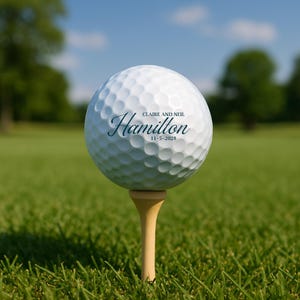 Puede incluir: Una pelota de golf blanca con el nombre "Hamilton" y la fecha "11-5-2028" impresa, colocada sobre un tee de madera. La pelota de golf est&aacute; en un campo de golf verde con un cielo azul y &aacute;rboles al fondo.