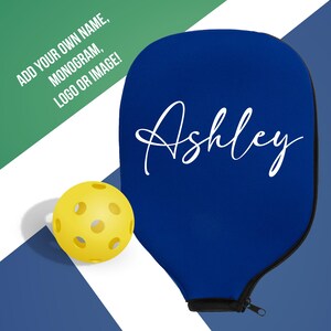 Pode incluir: Uma capa azul para raquete de pickleball com o nome "Ashley" em escrita branca. Uma bola de pickleball amarela est&aacute; ao lado da capa. O texto no canto superior esquerdo diz "ADICIONE SEU PR&Oacute;PRIO NOME, MONOGRAMA, LOGOTIPO OU IMAGEM!"