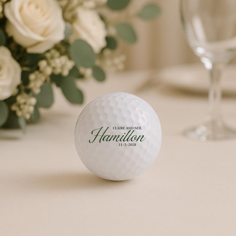 Puede incluir: Una pelota de golf blanca con el nombre "Hamilton" en letra cursiva y la fecha "11-5-2028". La pelota de golf est&aacute; sobre una superficie clara, con un fondo borroso de flores y una copa de vino.