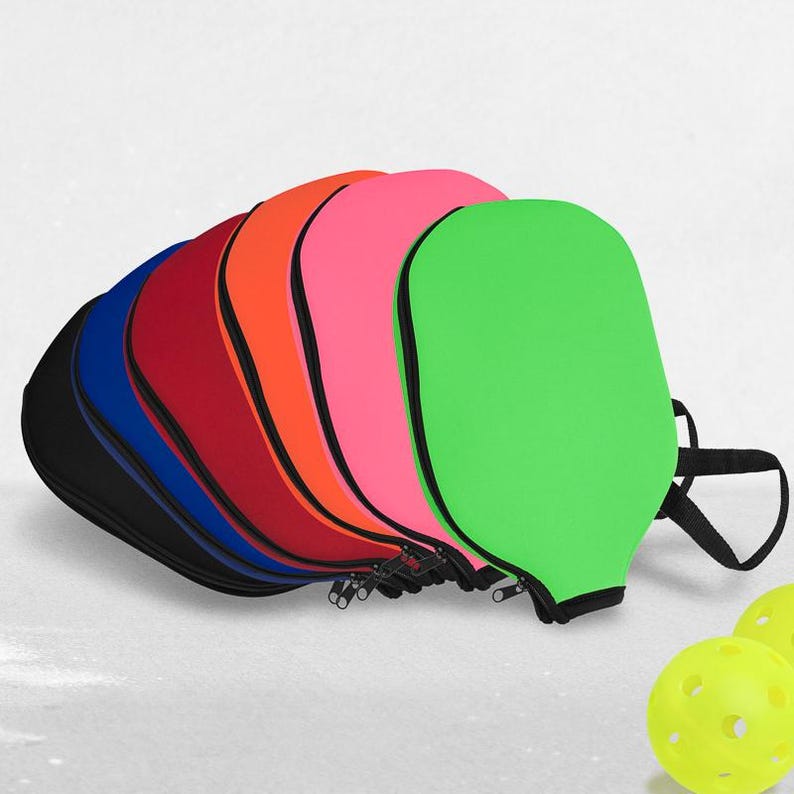 Pode incluir: Uma cole&ccedil;&atilde;o de capas coloridas para raquetes de pickleball em preto, azul, vermelho, laranja, rosa e verde. Cada capa tem um z&iacute;per preto e uma al&ccedil;a preta. Duas bolas de pickleball amarelas est&atilde;o em primeiro plano.
