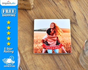 May include: A square ceramic coaster with a photo of a couple in a field. The man is carrying the woman on his back. They are both smiling. A red banner with the text "CHARLIE & DREW" is in front of them.