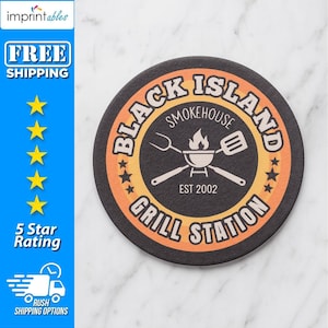 May include: A round black coaster with the text "Black Island Smokehouse Grill Station" in white and orange. The coaster features a grill graphic and the text "EST 2002".