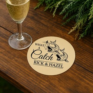 May include: A round, beige coaster with the text "WHAT A Catch" and "RICK & HAZEL" in black script. The coaster features a graphic of two fish. A glass of champagne is in the background.