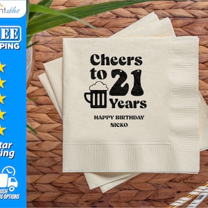May include: A stack of square, off-white paper napkins featuring black printed text that reads Cheers to 21 Years, with a beer mug graphic, and the text HAPPY BIRTHDAY NICKO printed in a smaller font below the main design.