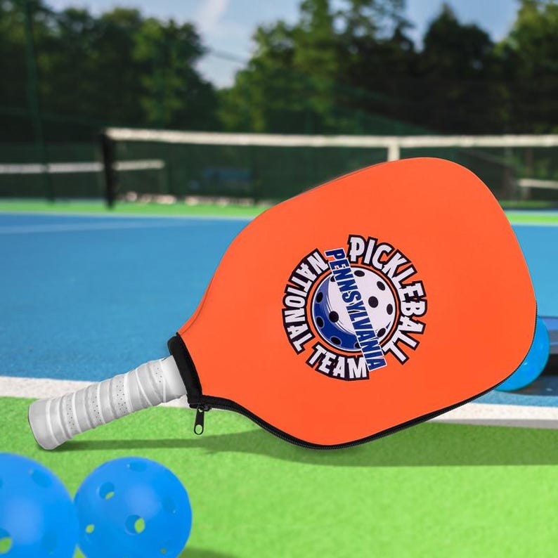 Pode incluir: Uma raquete de pickleball laranja com uma pega branca e o texto "PICKLEBALL PENNSYLVANIA NATIONAL TEAM" num design circular. Duas bolas de pickleball azuis est&atilde;o em primeiro plano. A raquete est&aacute; num campo azul.