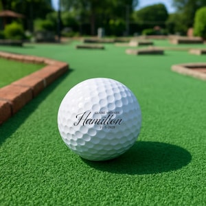 Puede incluir: Una pelota de golf blanca con el nombre "Hamilton" en escritura cursiva, los nombres "Claire y Neil" y la fecha "11-5-2028". La pelota est&aacute; sobre una superficie verde, posiblemente un campo de minigolf.