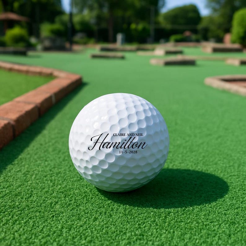 Puede incluir: Una pelota de golf blanca con el nombre "Hamilton" en escritura cursiva, los nombres "Claire y Neil" y la fecha "11-5-2028". La pelota est&aacute; sobre una superficie verde, posiblemente un campo de minigolf.