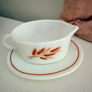 May include: A white milk glass gravy boat with a single handle and a pouring spout. The exterior features a brown wheat stalk pattern. It sits on a matching white saucer that has a thin brown circular line accenting the outer edge of the plate.