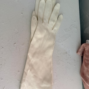 May include: A pair of long, off-white leather gloves featuring a smooth finish and three vertical stitched lines on the back of the hand. The gloves extend past the wrist, providing full coverage for the fingers and palms with a clean, simple design.