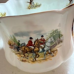May include: A white ceramic bowl with a scalloped rim and a gold-toned edge. The exterior features a colorful illustration of riders on horseback in red jackets with hunting dogs. The interior rim is decorated with small hunting dog motifs.