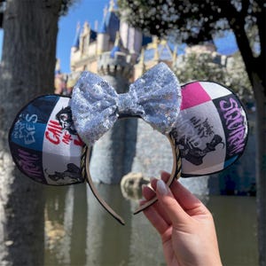 May include: A pair of mouse ears with a silver sequined bow. The ears have a black and white graphic design with text that reads "Call Me" and "You're in my world". The headband is gold and black.