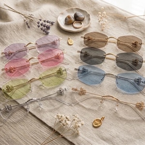 May include: Collection of rimless sunglasses with tinted lenses in various colors, including purple, pink, yellow, blue, and brown. The glasses have gold or silver frames and decorative accents. A small dish with stones and a ring, along with dried flowers, are also present.