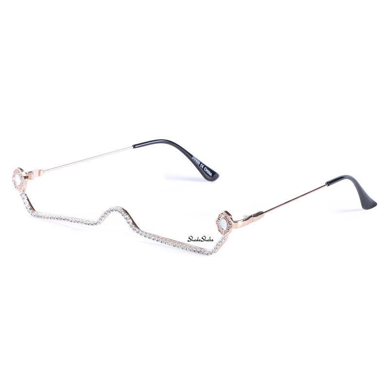 May include: Rose gold metal frame glasses with clear lenses and rhinestones along the frame. The glasses have a unique, curved shape.