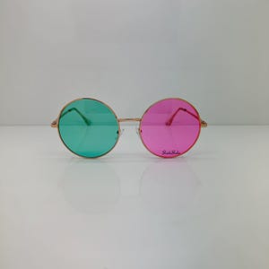 Op de afbeelding: Ronde zonnebril met gouden montuur en groene en roze glazen. Op het rechterglas staat de tekst "Shade Shade". Deze bril heeft een retro-stijl, die doet denken aan de jaren 60 en 70.