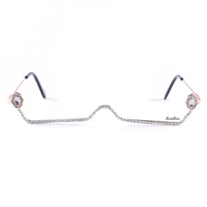 May include: A pair of rimless glasses with a gold frame and a row of rhinestones along the bottom. The glasses have a unique design with the rhinestones forming a wavy line.