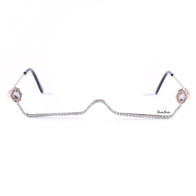 May include: A pair of rimless glasses with a gold frame and a row of rhinestones along the bottom. The glasses have a unique design with the rhinestones forming a wavy line.